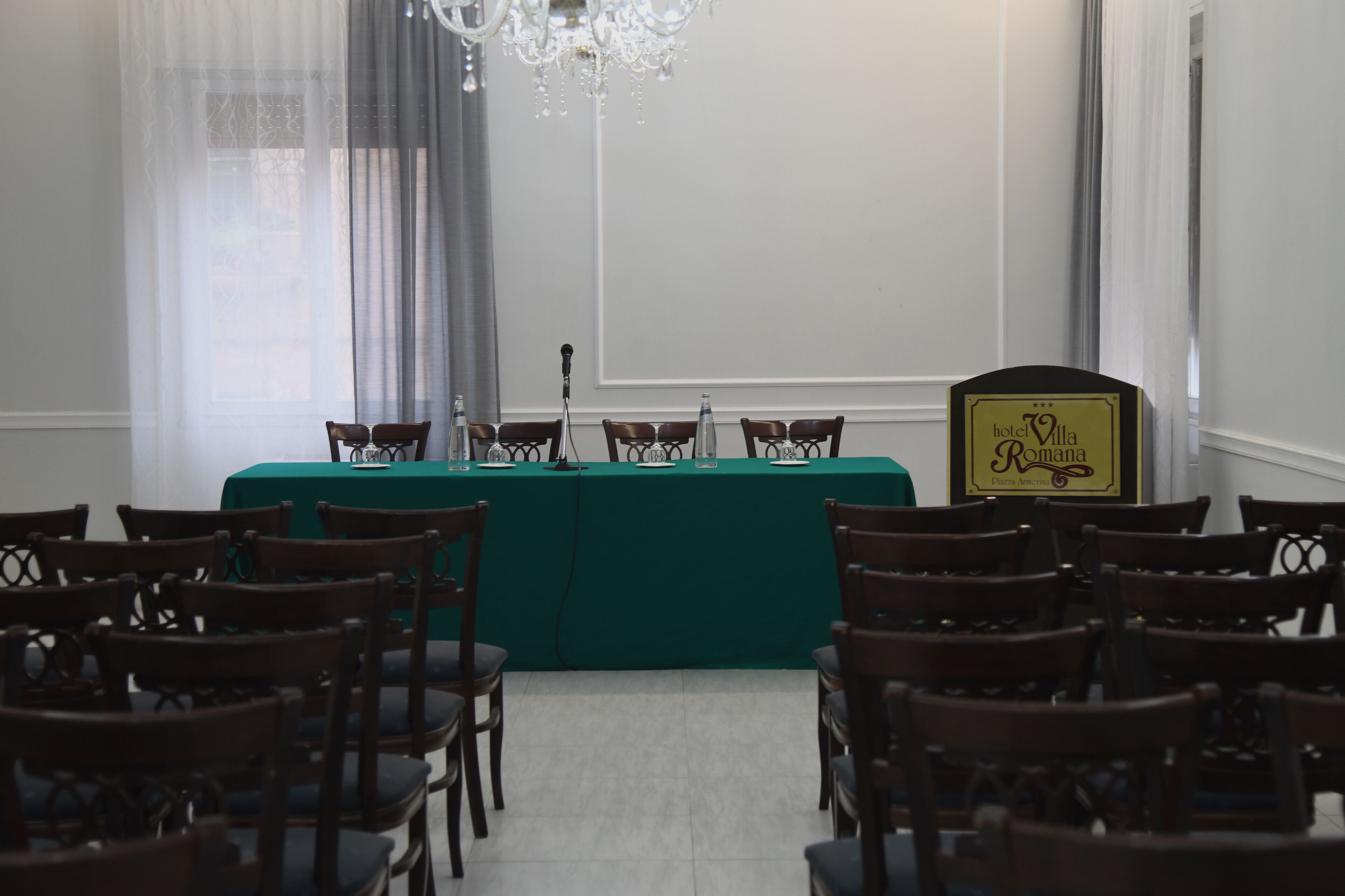 Una sala de conferencias con una mesa verde, sillas de madera, un soporte para micrófono y un letrero que dice 'Hotel Villa Romana'.