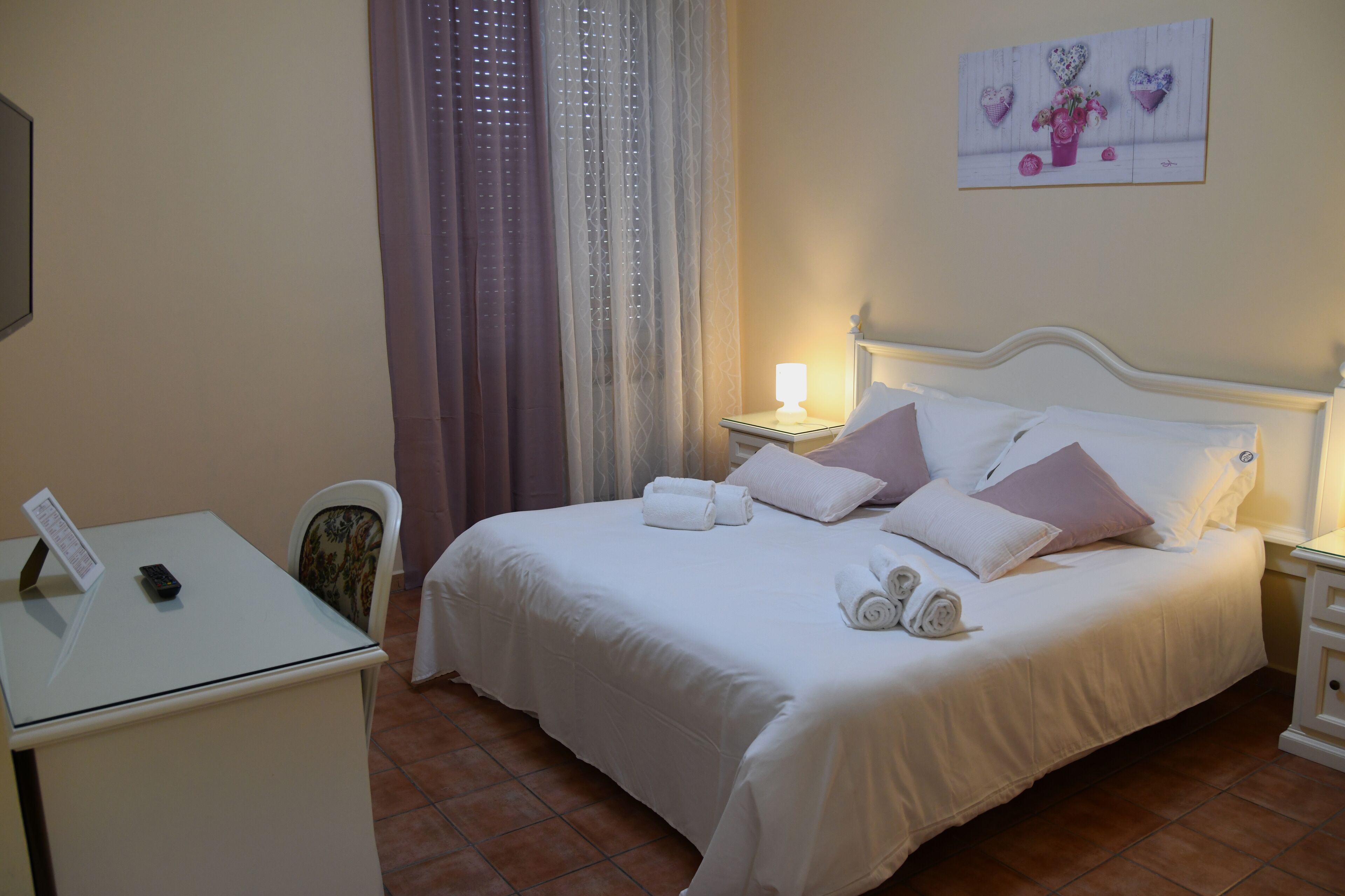 A neatly made bed with pillows and a rolled towel, a bedside table with a lamp, and a chair with a floral cushion.