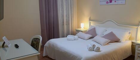 A neatly made bed with pillows and a rolled towel, a bedside table with a lamp, and a chair with a floral cushion.