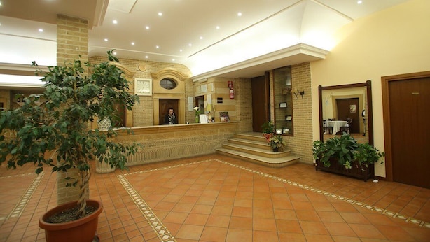 A lobby with a reception desk, a large potted plant, and a staircase leading to an upper level. - Hotel Villa Romana (Piazza Armerina)