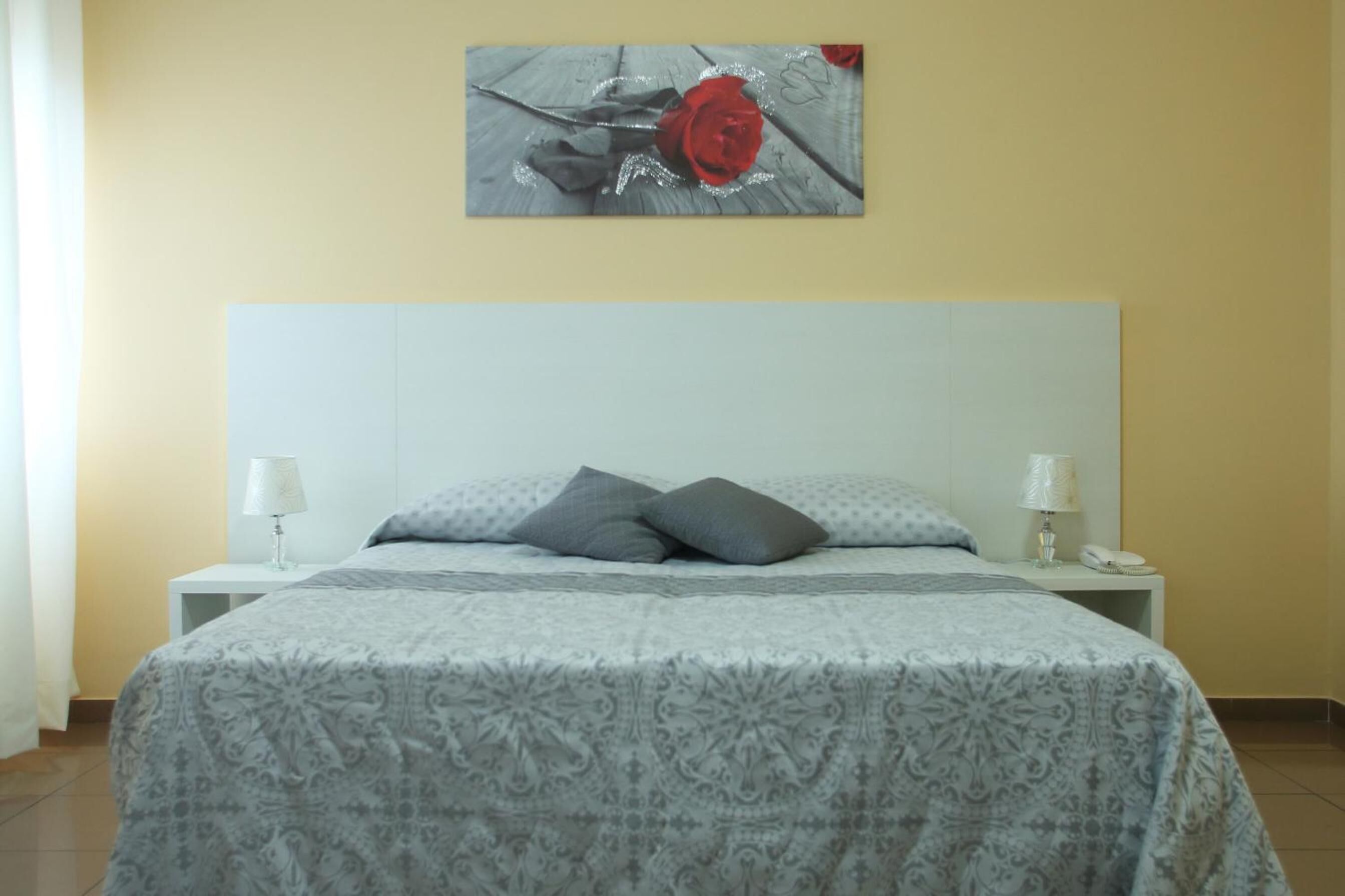 Een slaapkamer met een bed, twee nachtkastjes, een lamp op elk en een aan de muur hangend schilderij van een rode roos.