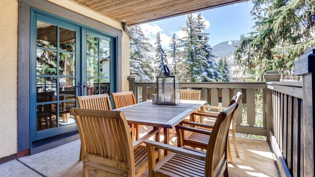 A covered patio with a wooden table and chairs, a glass door leading to a library, and a view of snow-covered trees and mountains. - 1-#28 One Willow Bridge 3BD (Vail)