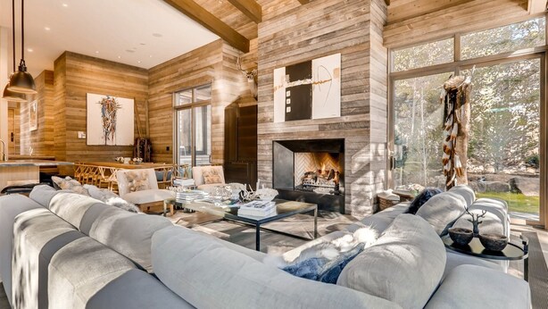 A modern log cabin interior with a large fireplace, a cozy seating area, and large windows offering a view of the outdoors. - 1-184 West Meadow Drive (Vail)