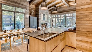 A modern kitchen with wooden cabinetry, a large island with a sink, pendant lights, and large windows offering views of the outdoors. - 1-184 West Meadow Drive (Vail)