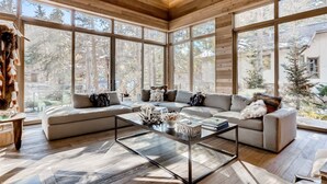 A spacious living room with a large sectional sofa, a glass coffee table, and a view of a forest through large windows. - 1-184 West Meadow Drive (Vail)
