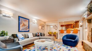 Ein modernes Wohnzimmer mit Sofa, Sessel und Couchtisch, ein Essbereich mit Stühlen und eine Küche mit orangefarbenen Lampen.