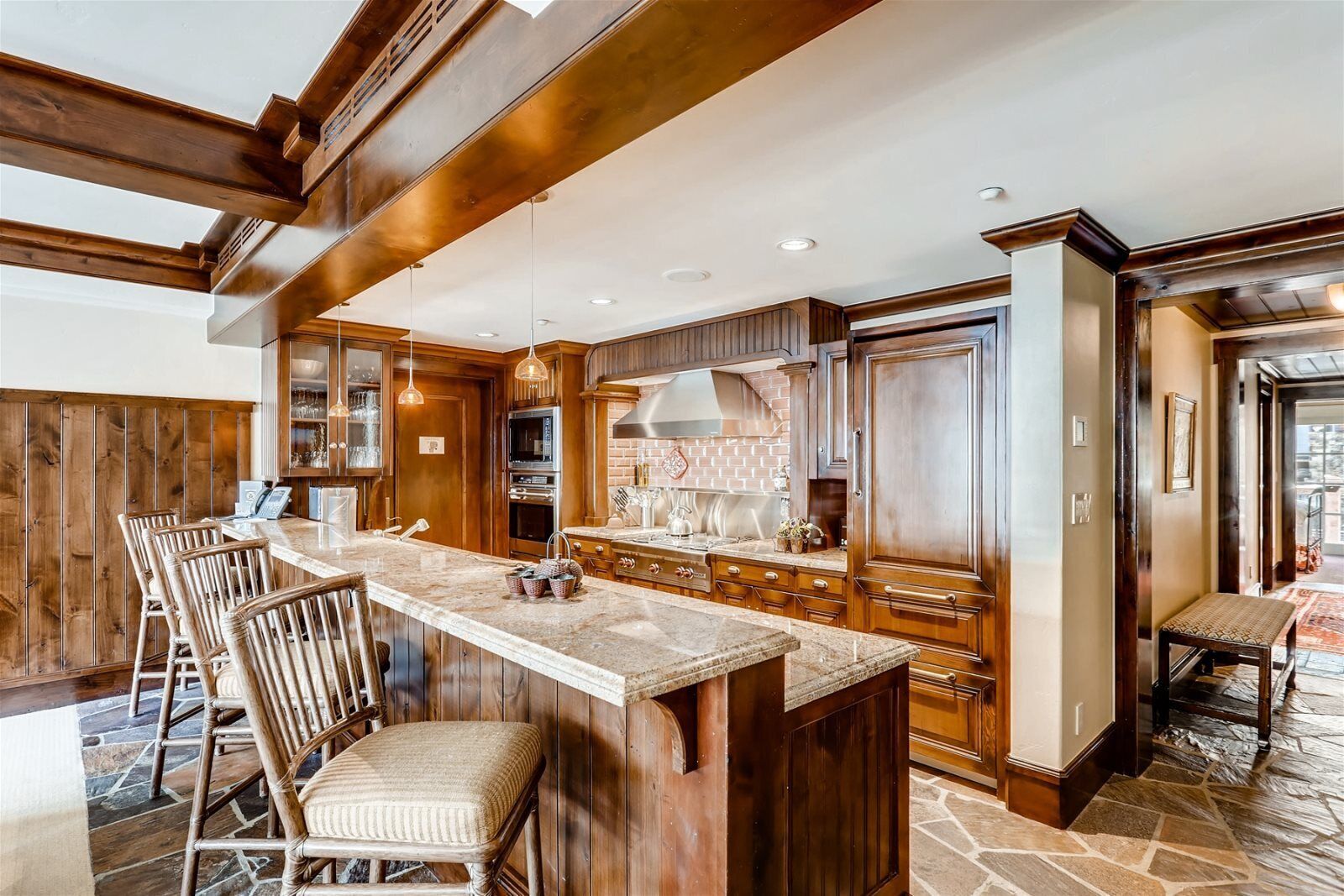 A spacious kitchen with a central island, wooden cabinetry, and a stone-tiled floor.