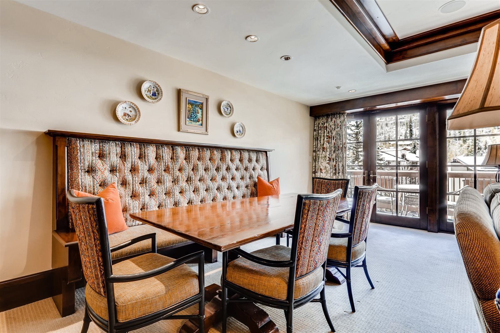 A dining area with a wooden table and upholstered chairs, a decorative wall panel, and a large window with a view of the outdoors.