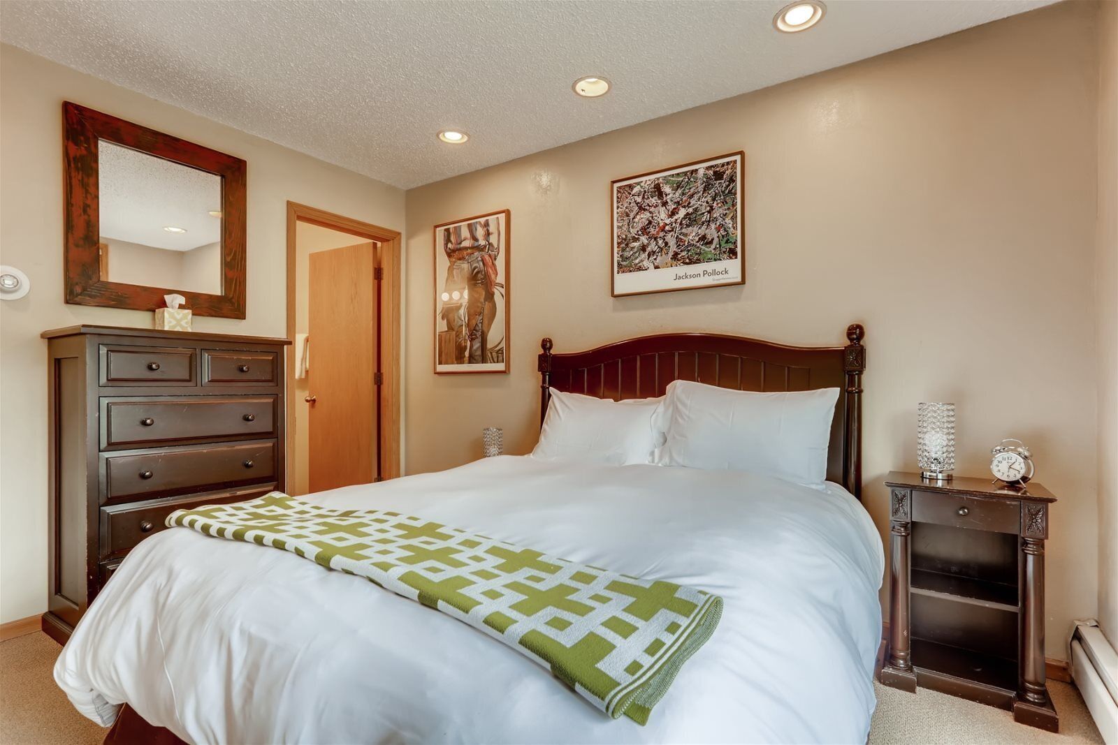 A bedroom with a large bed, a dresser, a nightstand, a mirror, and two framed artworks on the wall.
