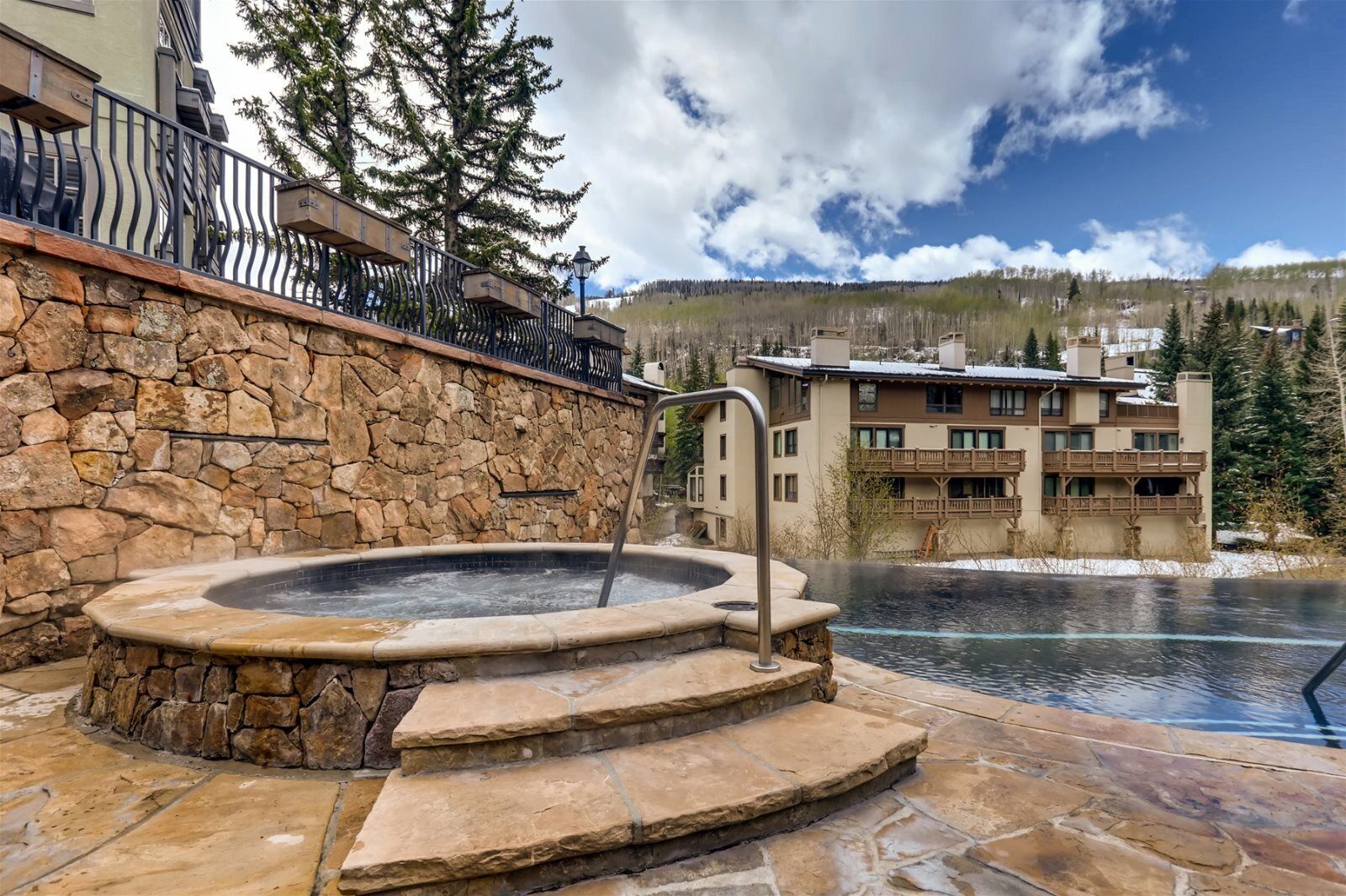 Eine Steinsichtmauer mit Whirlpool, Treppen, die zu einem Pool führen, und ein Gebäude mit Balkonen im Hintergrund.