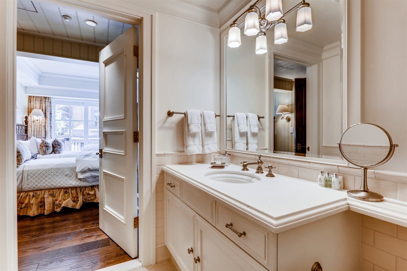 A hotel bathroom with a large mirror, a sink, and a bedside table.