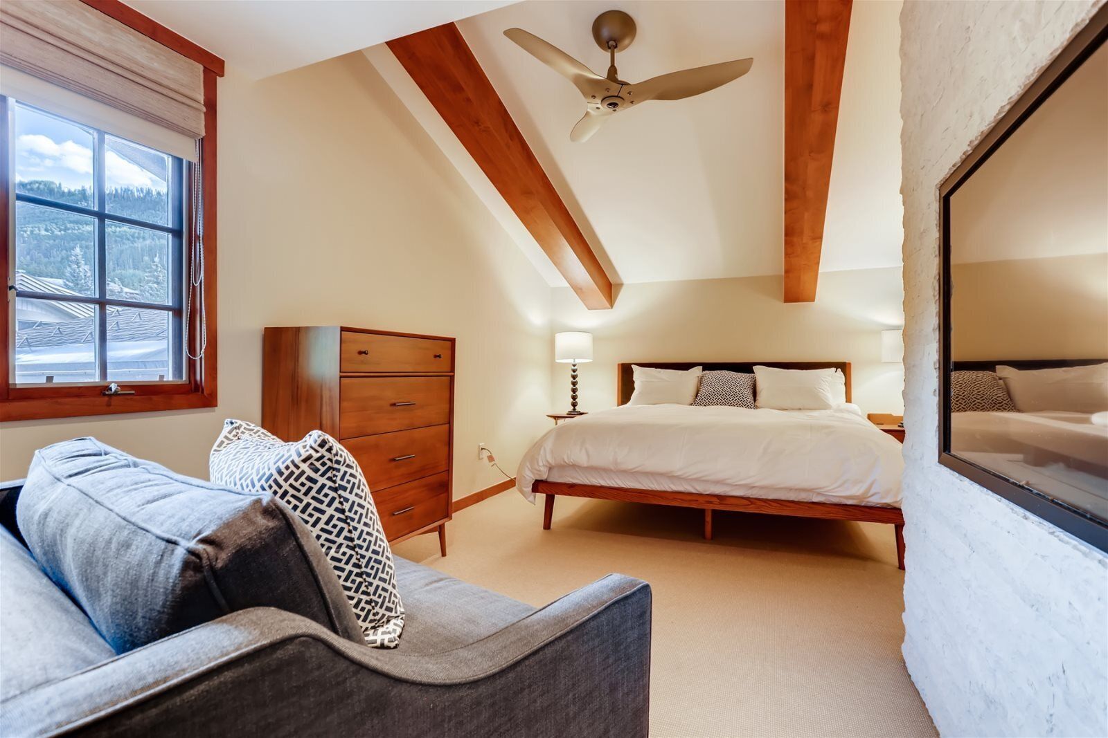 Une chambre à coucher comprenant un lit, une commode, un ventilateur de plafond et une fenêtre donnant sur des arbres.