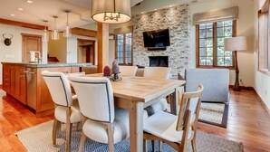 A spacious dining area with a wooden table and chairs, a stone accent wall, and a television mounted above a fireplace. - 1-385 Gore Creek Dr. Vorlaufer #303 (Vail)