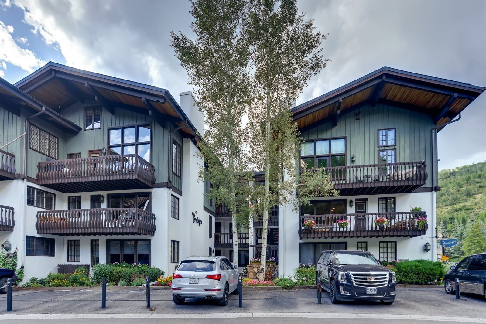 Un hotel de varios pisos con balcones, un área de estacionamiento y montañas al fondo.