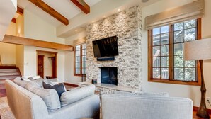 A spacious living room with a stone fireplace, a large TV mounted on the wall, and a comfortable seating area with a sofa and armchairs. - 1-385 Gore Creek Dr. Vorlaufer #303 (Vail)