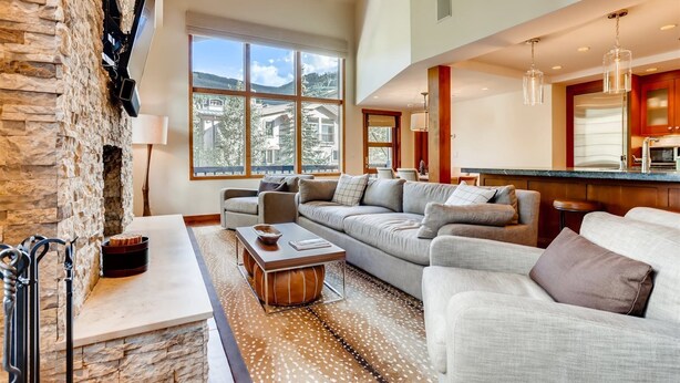 A spacious living room with a stone accent wall, large windows offering a view of a mountainous landscape, a cozy seating area with a fireplace, and a modern kitchen with wooden cabinets. - 1-385 Gore Creek Dr. Vorlaufer #303 (Vail)