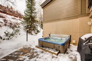 Uma banheira de hidromassagem em área externa nevada com um deck de madeira e uma casa ao fundo.
