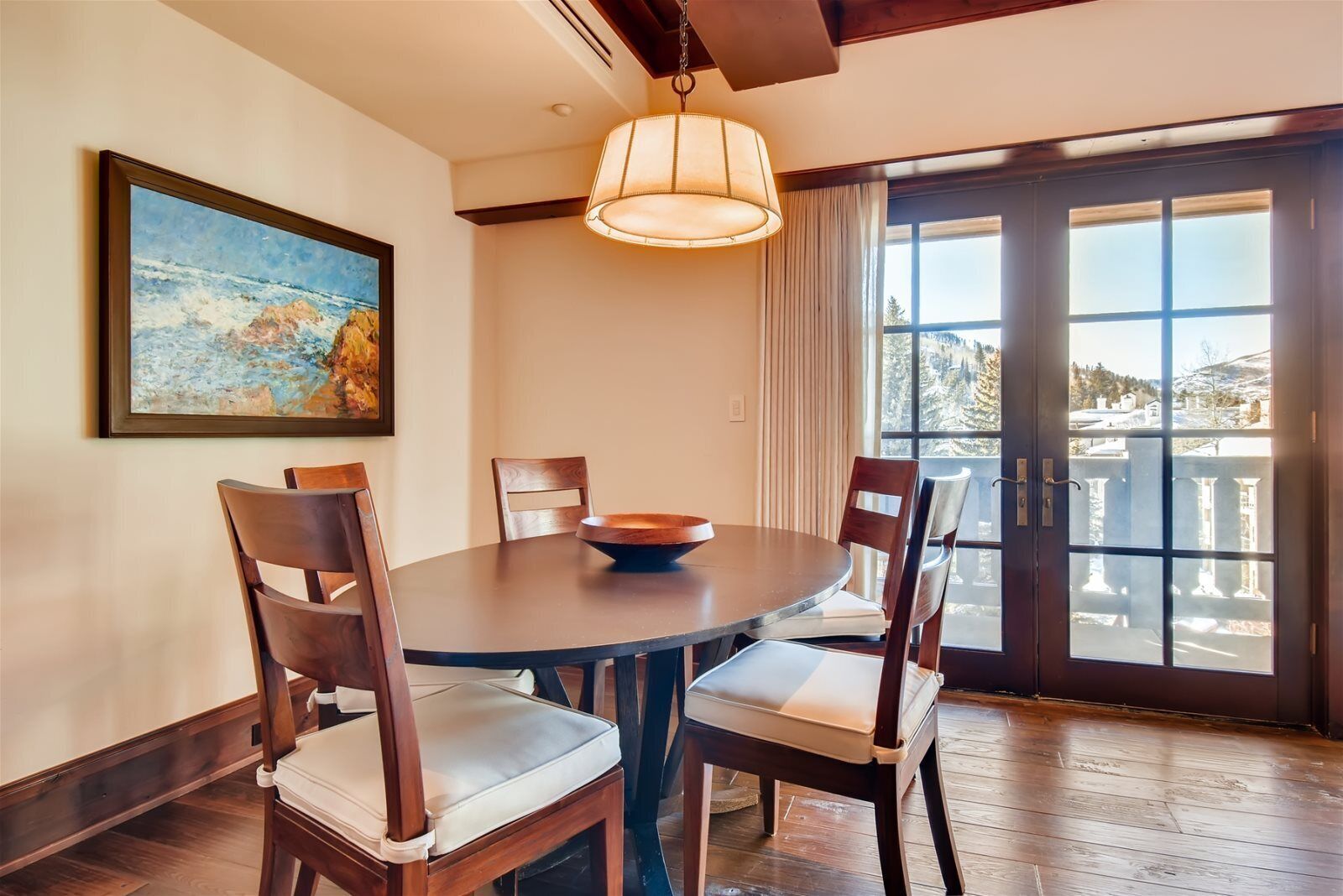 A dining area with a round table, four wooden chairs, a painting on the wall, and a large window with a view of a snowy landscape.