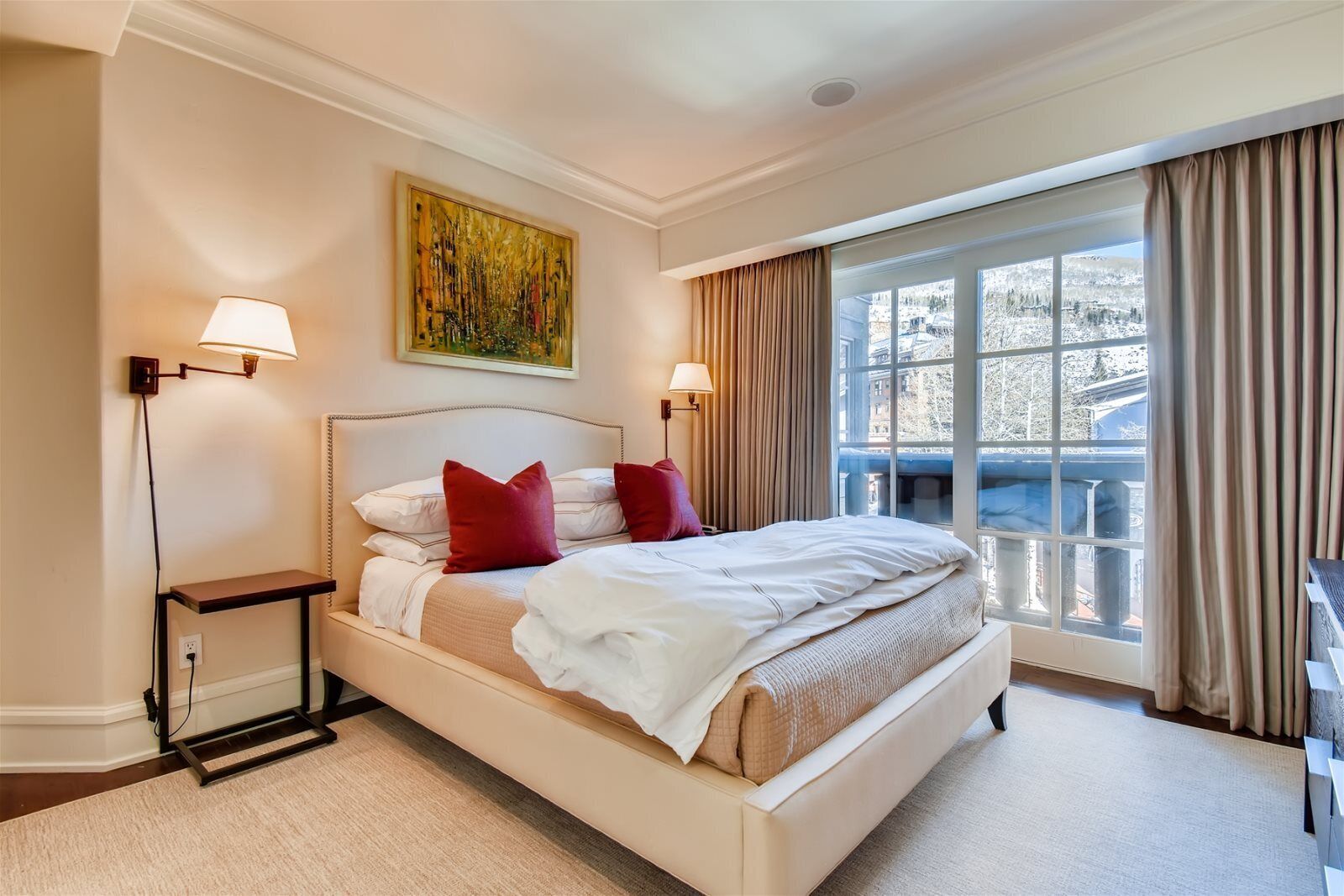 A hotel room with a large bed, two bedside lamps, a painting on the wall, and a view through a large window.