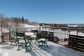 Un balcó amb cadires i taula coberts de neu, amb vista a un paisatge nevà i arbres allunyats.