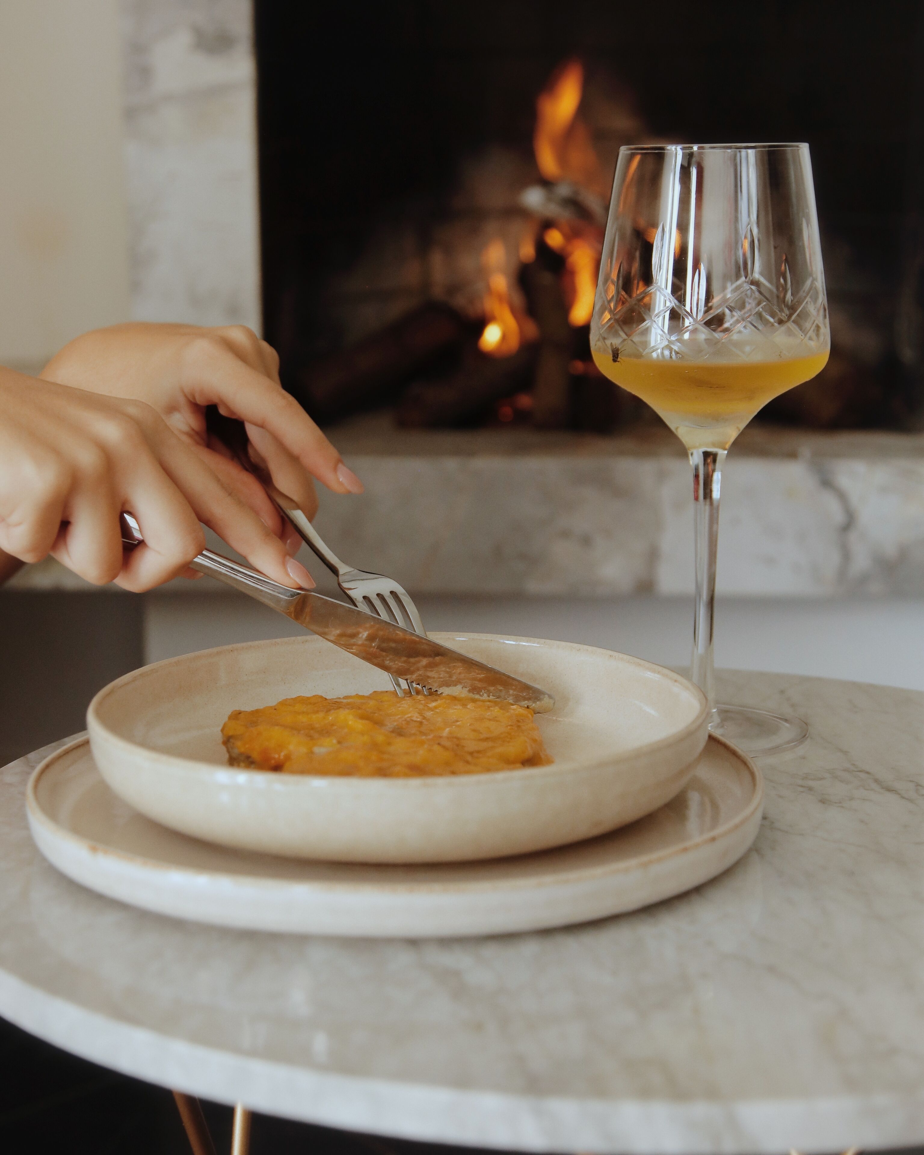 A plate with food and a glass of wine on a marble table with a fireplace in the background.