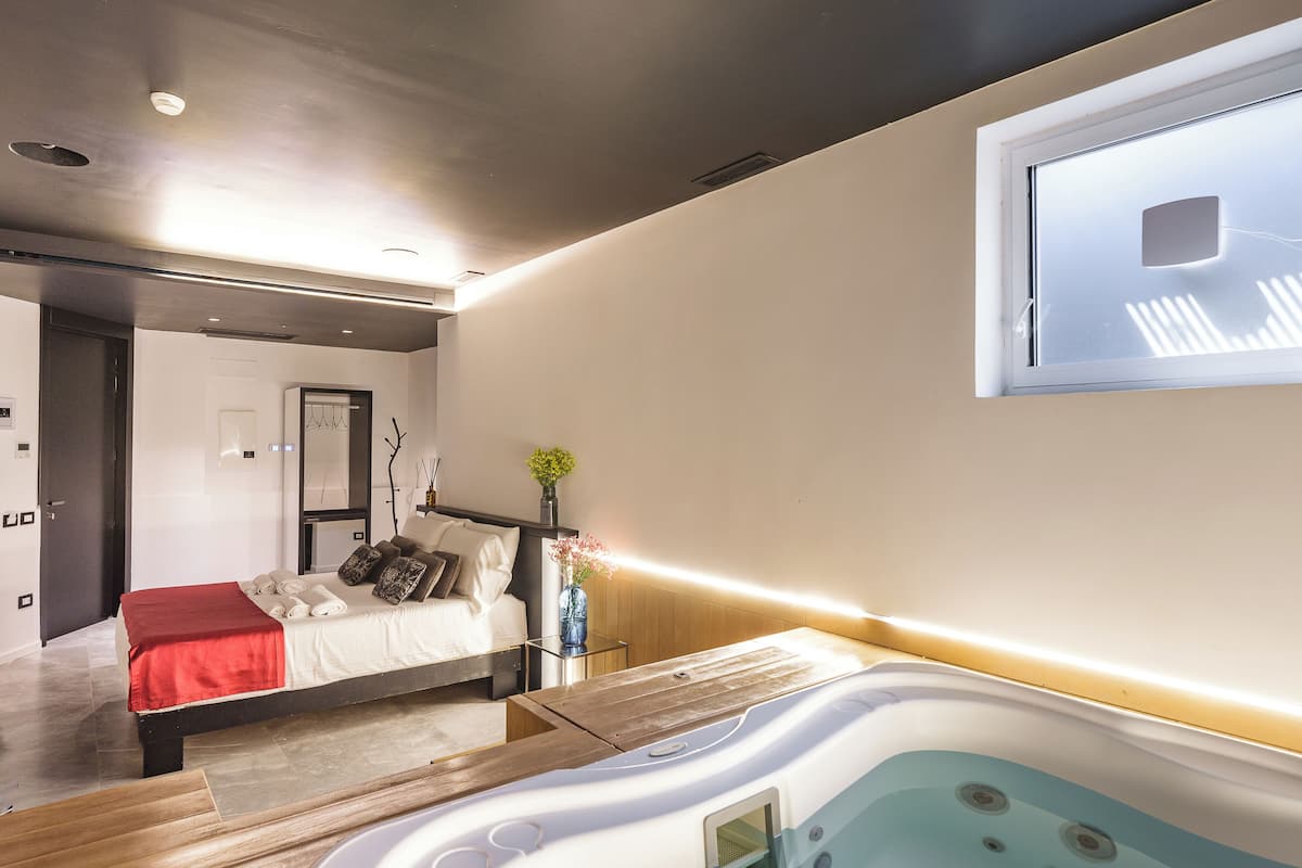 A modern bedroom with a bed, a bedside table, a nightstand, a large window, and a hot tub.