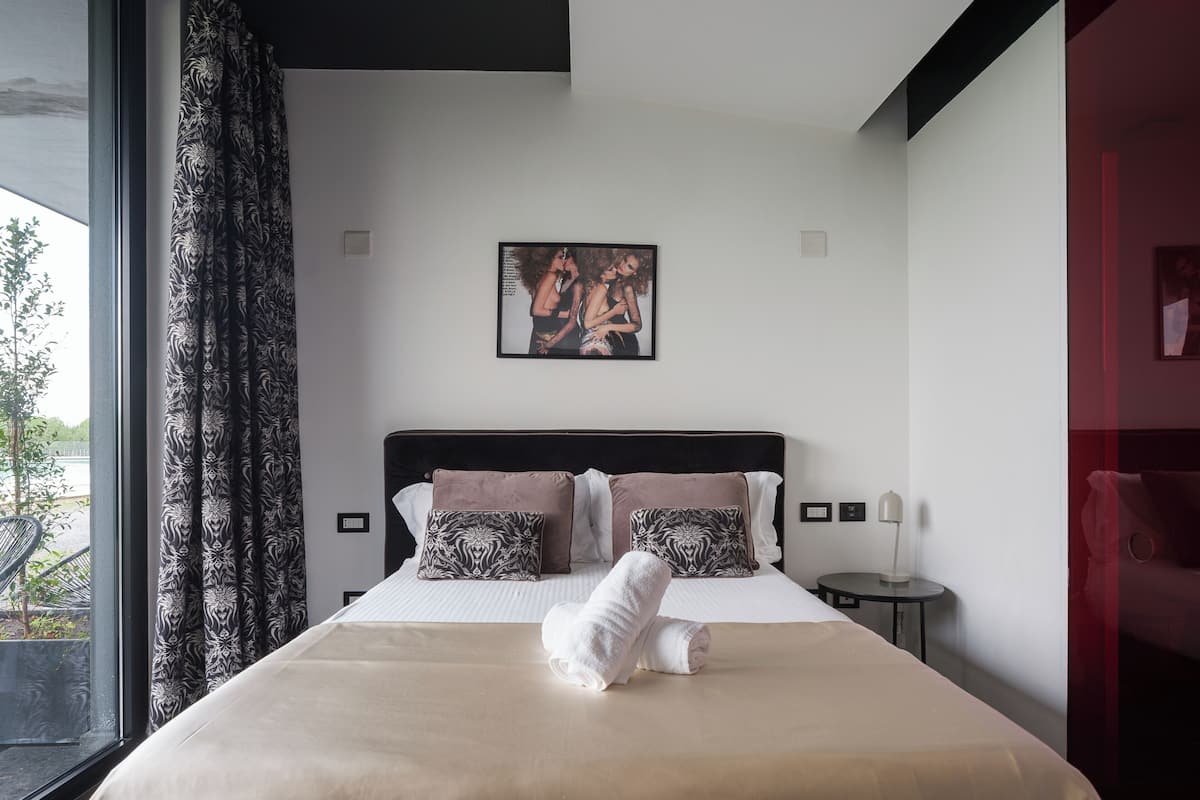 A neatly made bed with a white and beige bedspread, two patterned pillows, and a rolled white towel. A framed artwork hangs above the bed, and a window with curtains is visible to the left.
