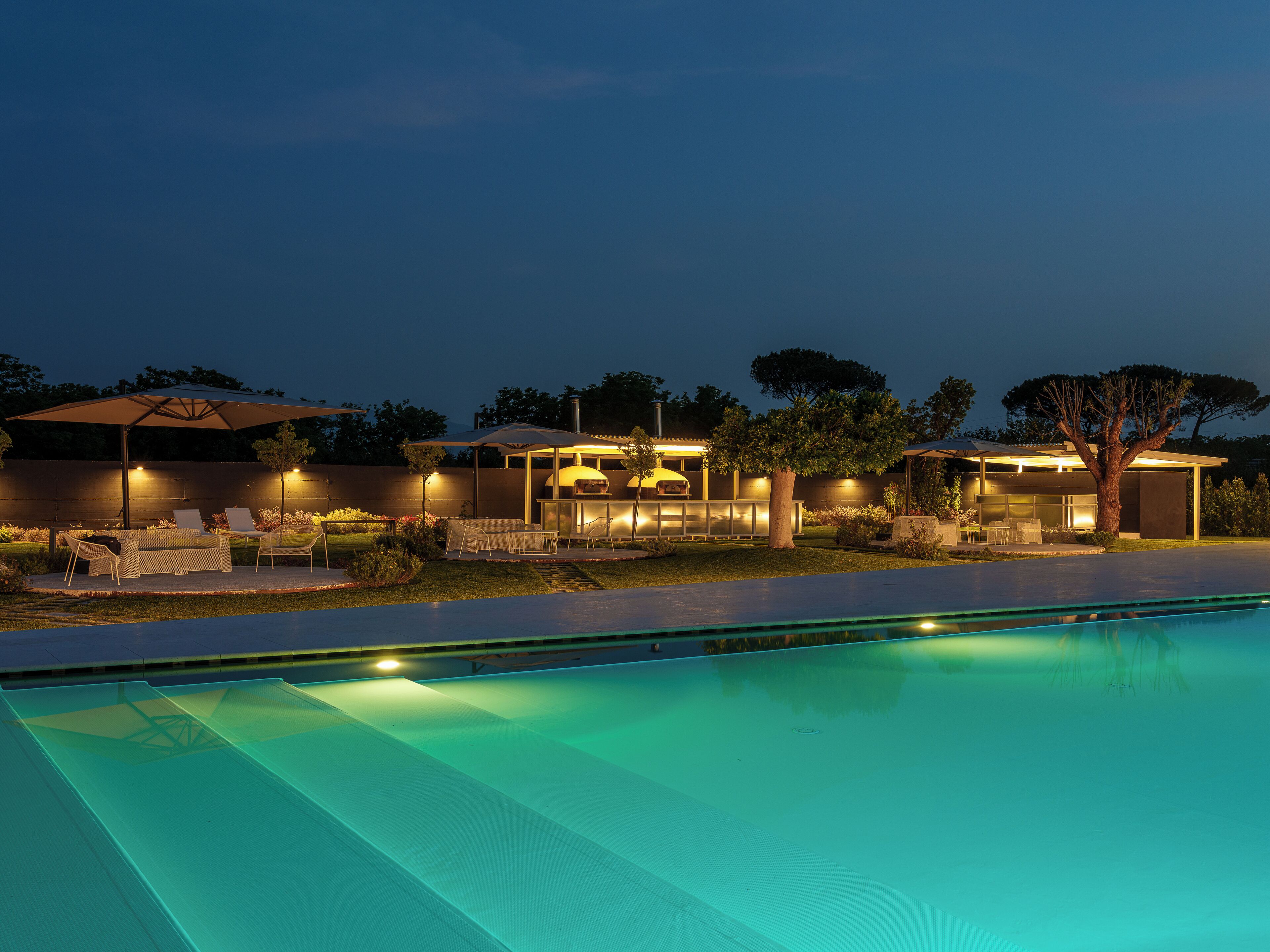 Zona junto a la piscina con mobiliario exterior, una barbacoa y una casa moderna al fondo.
