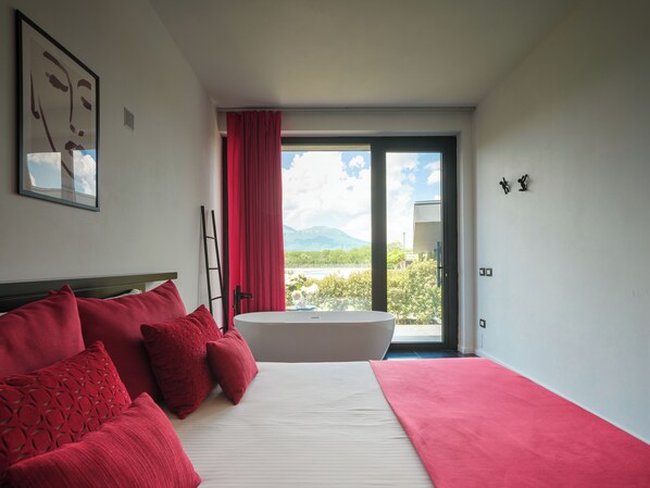 A bedroom with a large bed, red bedding, a bathtub, and a view of mountains and greenery. - Secret Garden (Palma Campania)