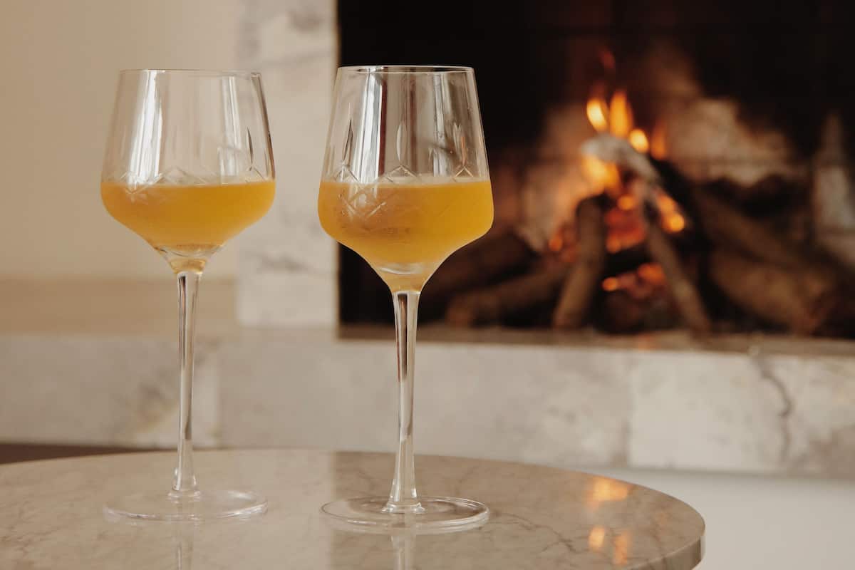 Two glasses of orange juice on a marble table with a fireplace in the background.