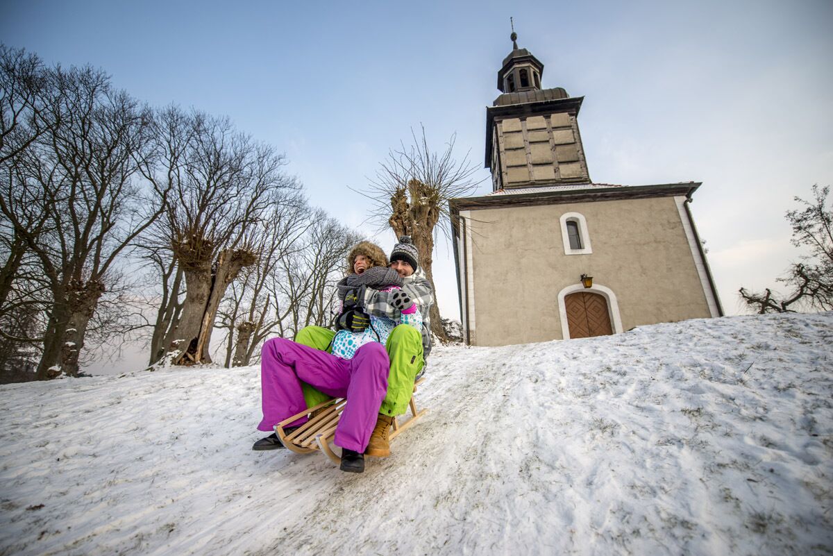 Two people sledding down a snowy slope with a church in the background.