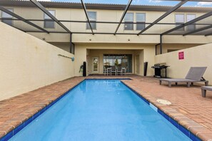 Une piscine bordée de carreaux, un patio couvert avec des chaises, et un bâtiment avec des fenêtres et une porte en arrière-plan.