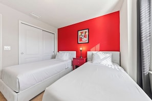 Habitación de hotel con dos camas, una pared de acento roja, un cuadro enmarcado, una mesita de noche con lámpara y una puerta.