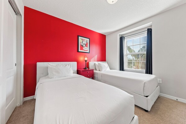 A room with two beds, a red accent wall, a window with blinds, and a framed picture on the wall.