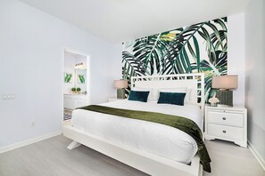 Une chambre à coucher comprenant un lit, des tables de chevet, un mur orné d’un motif de feuilles tropicales et une porte donnant accès à une salle de bain.