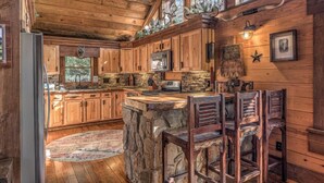 A rustic kitchen with wooden cabinets, a stone island, and a bar area with wooden chairs. - Winding Log Ridge- Pet Friendly | Screened Porch | 11 miles from Downtown (Blue Ridge)