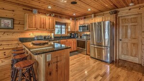 A wooden cabin kitchen with a central island, bar stools, stainless steel appliances, and wooden cabinets. - Tranquility- Pet Friendly | Mountain View | Hot Tub | Pool Table | Shuffleboard | Ping Pong (Mineral Bluff)