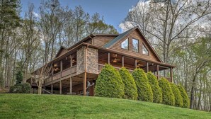A wooden cabin with a stone foundation, large windows, and a balcony. - Tranquility- Pet Friendly | Mountain View | Hot Tub | Pool Table | Shuffleboard | Ping Pong (Mineral Bluff)