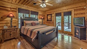 A wooden cabin bedroom with a bed, two bedside lamps, a ceiling fan, a TV, and a wooden cabinet. - Tranquility- Pet Friendly | Mountain View | Hot Tub | Pool Table | Shuffleboard | Ping Pong (Mineral Bluff)
