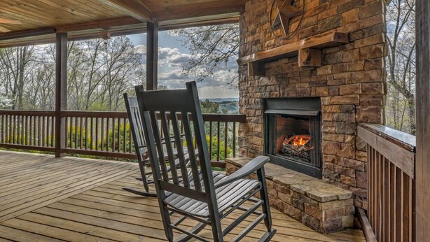 A wooden deck with a stone fireplace and a wooden chair. - Tranquility- Pet Friendly | Mountain View | Hot Tub | Pool Table | Shuffleboard | Ping Pong (Mineral Bluff)
