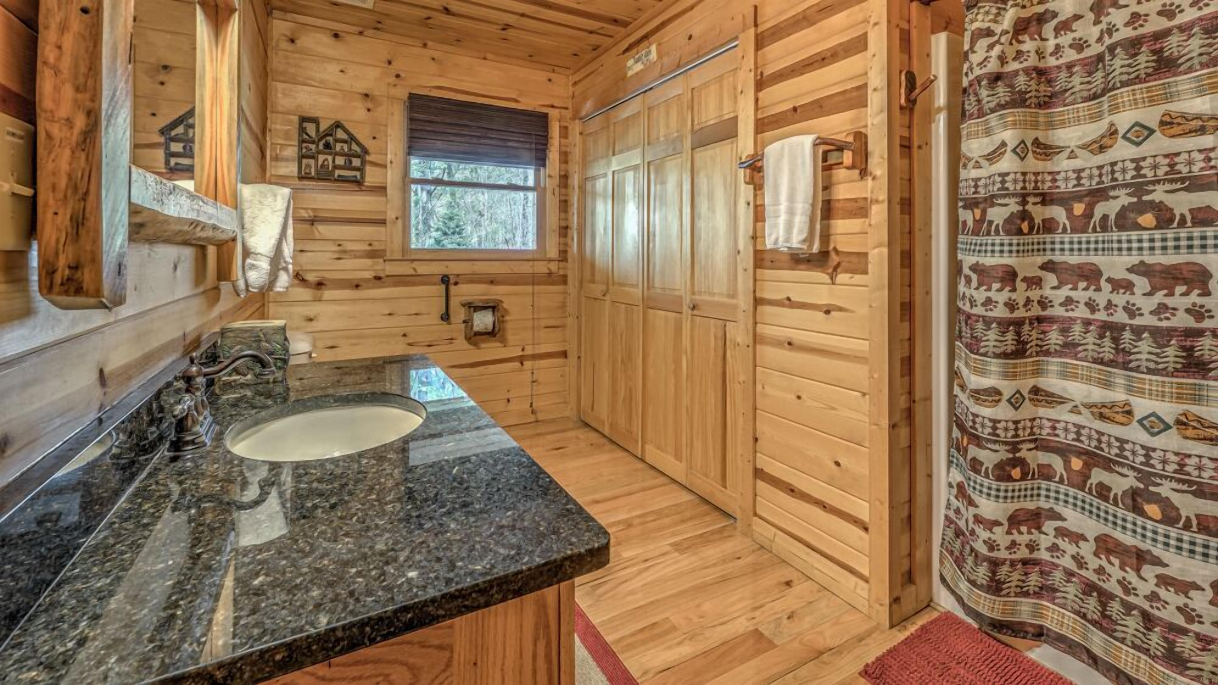 A wooden cabin bathroom with a granite countertop sink, wooden walls, a window, and a shower curtain with animal prints.