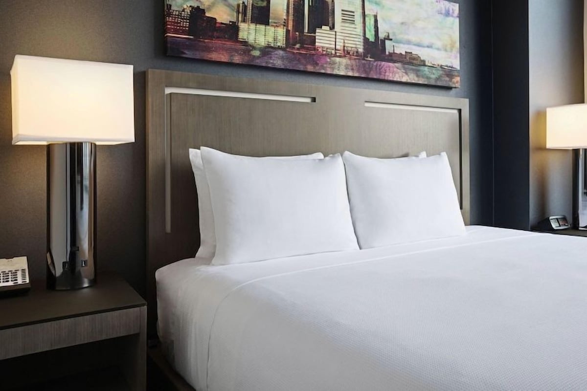 A hotel room with a large bed, two bedside lamps, a nightstand with a phone, and a cityscape painting on the wall.