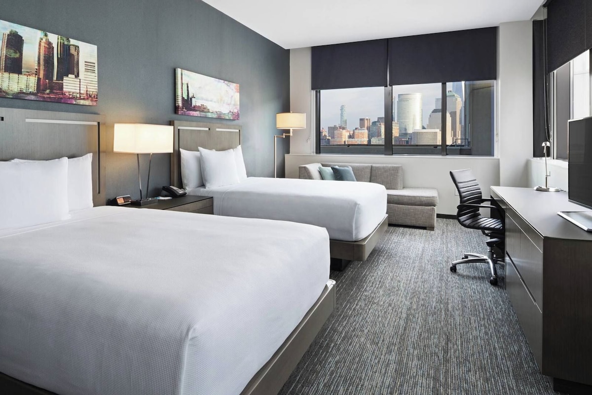 A hotel room with two beds, a desk, a chair, a lamp, and a large window with a city view.