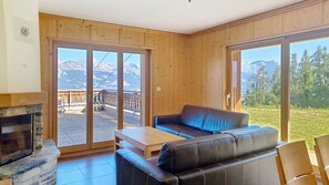 Ein Zimmer mit Kamin, einer Couch, einem Tisch und großen Schiebeglastüren, die auf eine Terrasse mit Bergblick führen.