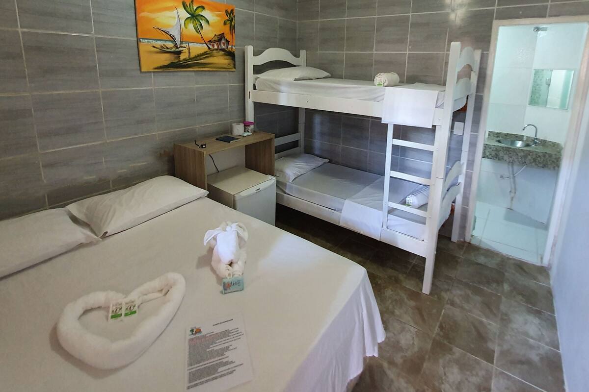 Habitación de hotel con literas, una mesita de noche con lámpara, un escritorio pequeño con silla y un baño con lavamanos y espejo.
