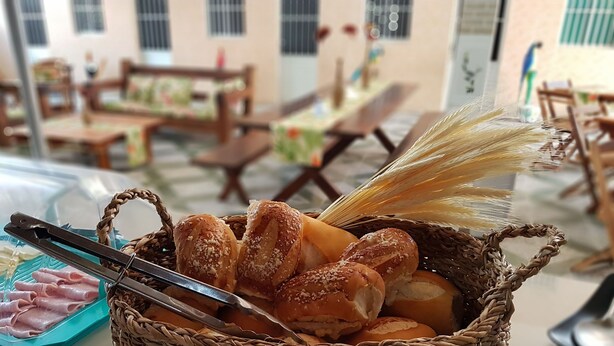 Uma cesta de pão e presunto sobre uma mesa em área de refeições, com mobiliário de madeira e uma janela ao fundo.