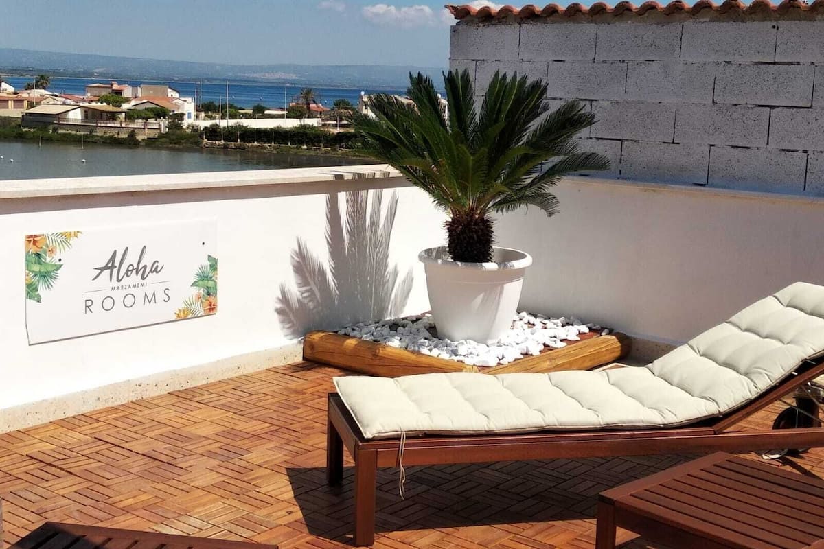 A balcony with a lounge chair, a potted plant, and a sign reading "Aloha Marzamemi Rooms".