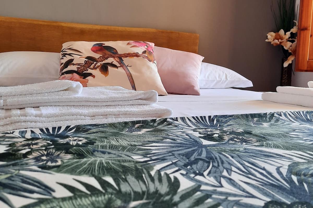 A neatly made bed with floral and solid pillows, a wooden headboard, and a tropical leaf-patterned bedspread.