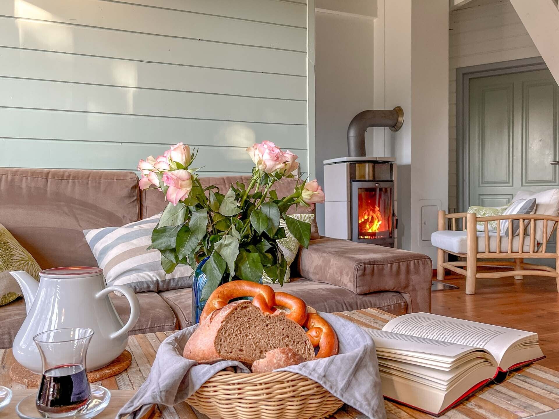 A cozy living room with a fireplace, a sofa, a chair, a vase with flowers, a basket of bread, a book, and a glass of drink.