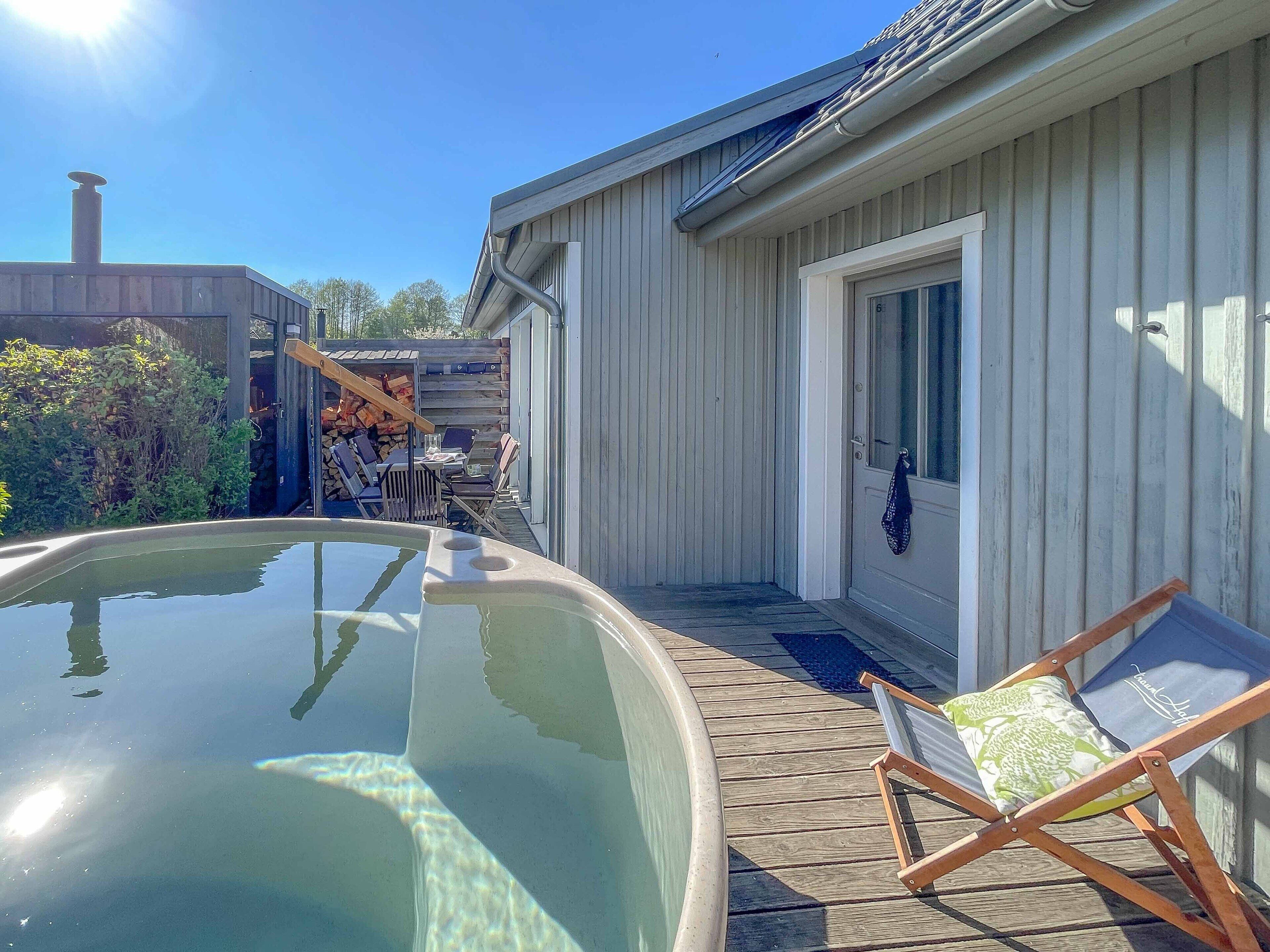 Une terrasse en bois avec un jacuzzi, un espace salon extérieur couvert et un bâtiment doté d’une porte et d’une fenêtre.